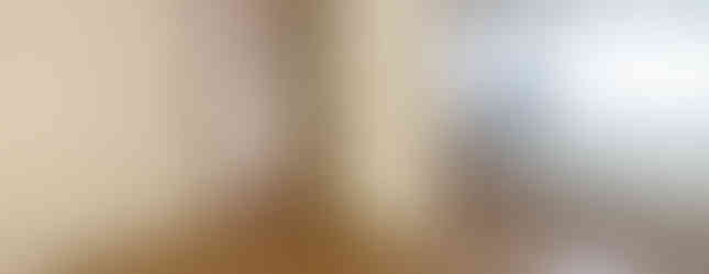 A photo of a spacious living room with a wooden floor and wallpapered walls. The room features a sliding door leading to another room, as well as a doorway to the kitchen. The kitchen is visible in the background with white cabinets and countertops.