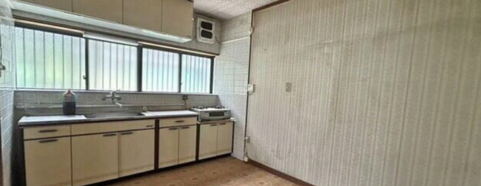 A dated kitchen with beige cabinets and a white countertop. A gas stove and a range hood are installed above the countertop. A sink with a faucet is located to the left of the stove. A large window with sliding glass doors overlooks the backyard. The walls are wallpapered and the floor is covered in patterned tiles.