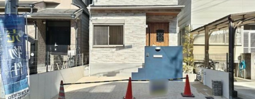 This image shows the exterior of a two-story house in Japan. The house has a gray exterior with a traditional Japanese-style roof. There is a small front yard with a tiled walkway and a driveway. The house is located on a street with other houses nearby.