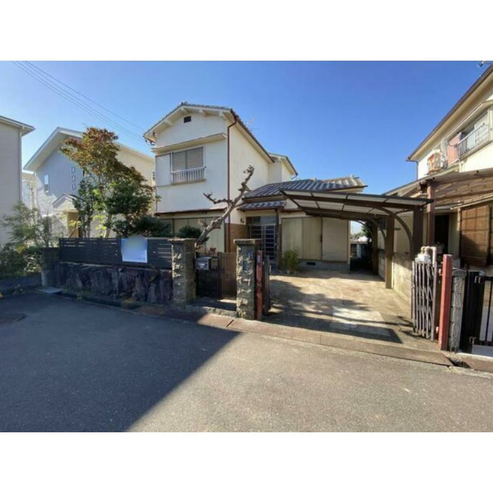 This photo shows the front exterior of a traditional Japanese house. The house is painted white with a dark brown roof and wooden accents. There is a small front porch with a pair of wooden doors. The house is surrounded by trees and greenery.