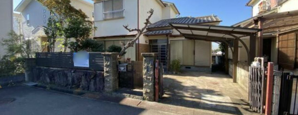 This photo shows the front exterior of a traditional Japanese house. The house is painted white with a dark brown roof and wooden accents. There is a small front porch with a pair of wooden doors. The house is surrounded by trees and greenery.