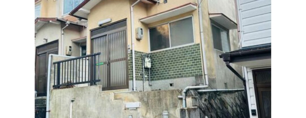 The photo shows a two-story house with a light-colored facade and a dark-colored roof. The house has several windows, a front door with a porch, and a driveway with a garage door.