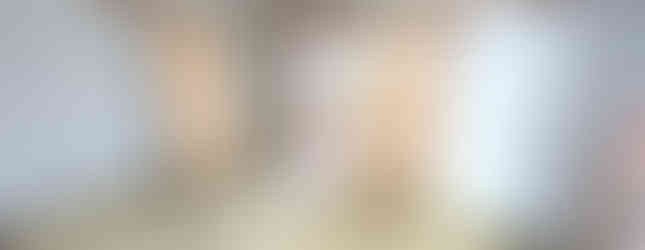 This photo shows the interior of a traditional Japanese-style room. The room features tatami mats, sliding paper doors (fusuma), and a ceiling with decorative beams. The walls are covered in wallpaper with a floral pattern. The floor is made of wood and has a parquet pattern. There is a window on the right side of the room, allowing natural light to enter. The room is currently empty except for some furniture and boxes.