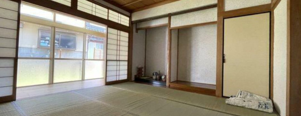A traditional Japanese room with tatami mat flooring and multiple shoji screen doors, some of which are open to reveal a view of a garden. A raised platform (tokonoma) with a small display is present in the corner. The room has a wooden ceiling with exposed beams.
