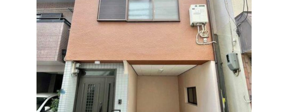 The image shows a two-story house with a brown facade. The house has a garage on the ground floor and a balcony on the second floor. There is a black door and a mailbox in front of the house. The house is located in a residential area with other houses visible in the background.