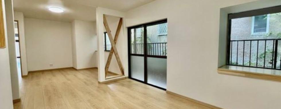 This photo shows a spacious living room with light wood flooring and white walls. There are two windows on the right side of the room, one with a view of a balcony and the other with a view of the street. A wooden partition separates the living area from the dining area.