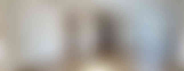 This photo shows a spacious and well-lit living room with a modern design. The room features wood-look laminate flooring, white walls, and a decorative ceiling with recessed lighting. A large window provides ample natural light. A sliding door leads to a separate room, and another doorway reveals a glimpse of a kitchen with white cabinets and appliances. The overall impression is one of a clean, open, and inviting space.