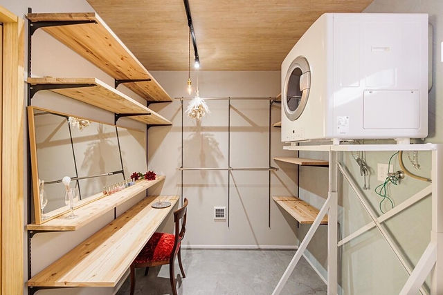 A bright and airy laundry room with wooden shelves and a dressing table. A washer and dryer are stacked in the corner.