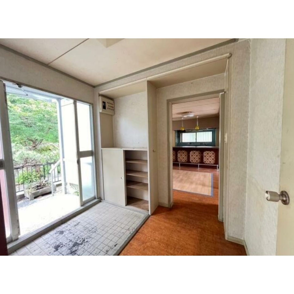 The photo shows an entryway with a tiled floor and a wooden door leading to the main living area. There are built-in shelves on one wall and a large window that looks out onto a garden. The overall impression is of a vintage, retro-style space.