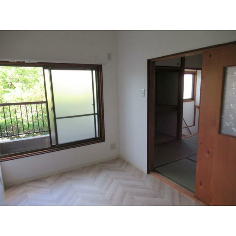 The photo shows a spacious room with a large window overlooking a green area. The room has a light wood floor and white walls, creating a bright and airy atmosphere. A sliding door leads to another room with traditional Japanese tatami mats. The overall impression is one of tranquility and openness.