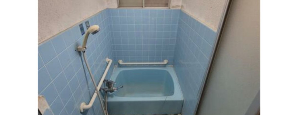 The photo shows a small bathroom with blue tiled walls and floor. There is a bathtub with a shower head, a toilet, and a sink. The bathroom appears to be outdated and in need of some repairs.
