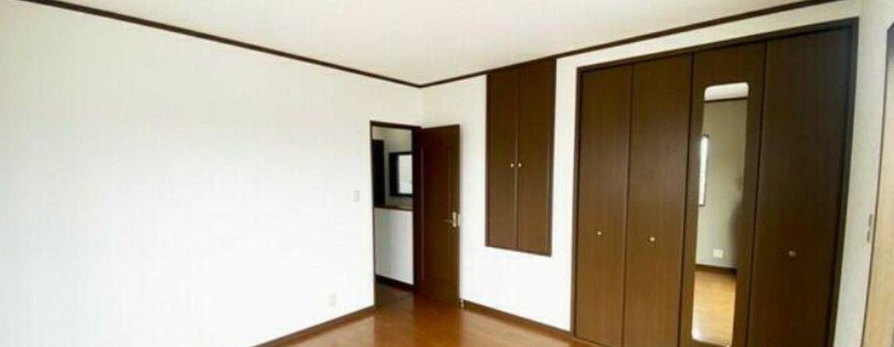 This image shows the interior of a bedroom. The room features a wooden floor, white walls, and a large closet with mirrored doors. There is also a door leading to another room.