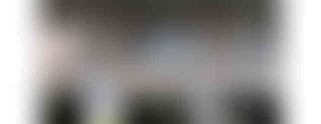 A three-story brick townhouse with a balcony on each floor. The first floor has a garage door and a small front porch. There are laundry lines hanging from the balconies on the second and third floors. The sky is blue with some clouds.