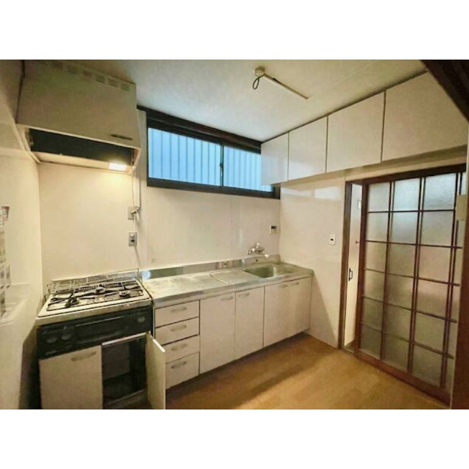 The photo shows a small, clean kitchen with white cabinets and a stainless steel countertop. A gas stove with a black hood is on the left side of the counter. There is a double sink with a faucet in the center of the counter. A sliding glass door with a frosted glass panel is on the right side of the room, leading to another room. A small window with blinds is above the counter, providing natural light.
