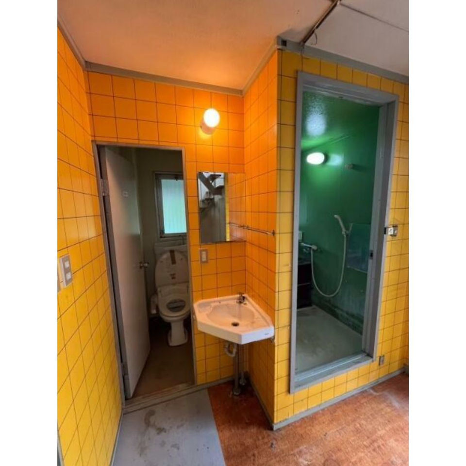The photo shows a vintage-style bathroom with bright yellow tiled walls. There is a sink, a toilet, and a shower stall with a green tiled wall. The overall impression is of a unique and nostalgic space.