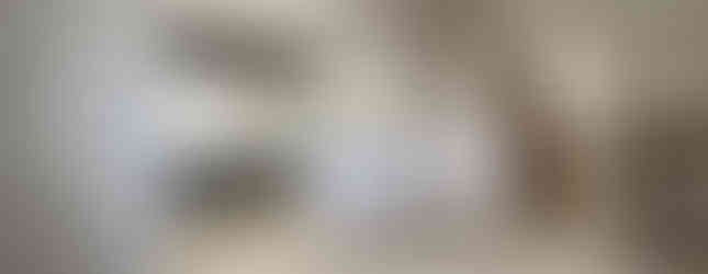 The photo shows a kitchen and living room area in a modern Japanese home. The kitchen features dark gray cabinets and a white countertop. A breakfast bar separates the kitchen from the living room. The living room has a light wood floor and a door leading to another room. The overall impression is of a clean, modern, and spacious living area.