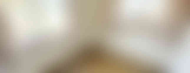 A small bedroom with light-colored walls and hardwood floors. Two windows with curtains are located on opposite sides of the room, providing natural light. The room is empty, with no furniture present.