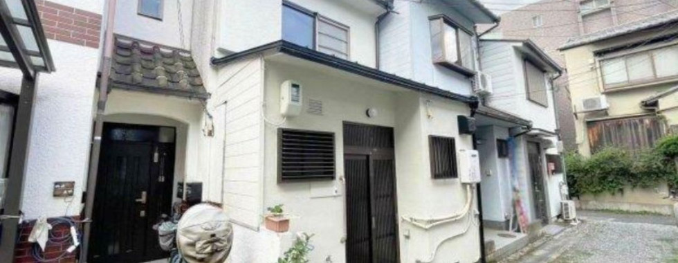 This image shows the exterior of a Japanese house. The house has a white exterior with a traditional Japanese-style roof. There is a small front yard with a tiled walkway and a bicycle. The house is located on a narrow street with other houses nearby.