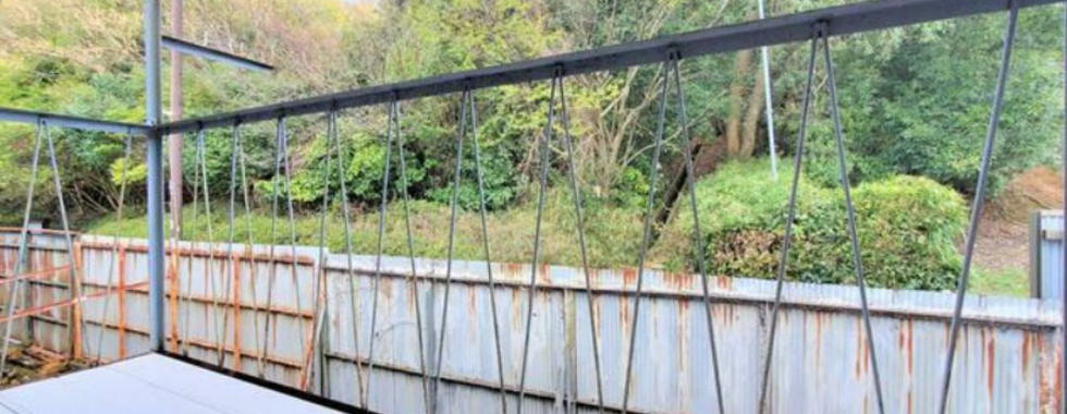 The photograph shows a balcony with a metal railing overlooking a green space. The balcony has a corrugated metal roof and a wooden floor. Beyond the railing, there's a view of trees, bushes, and a rusty corrugated metal fence.