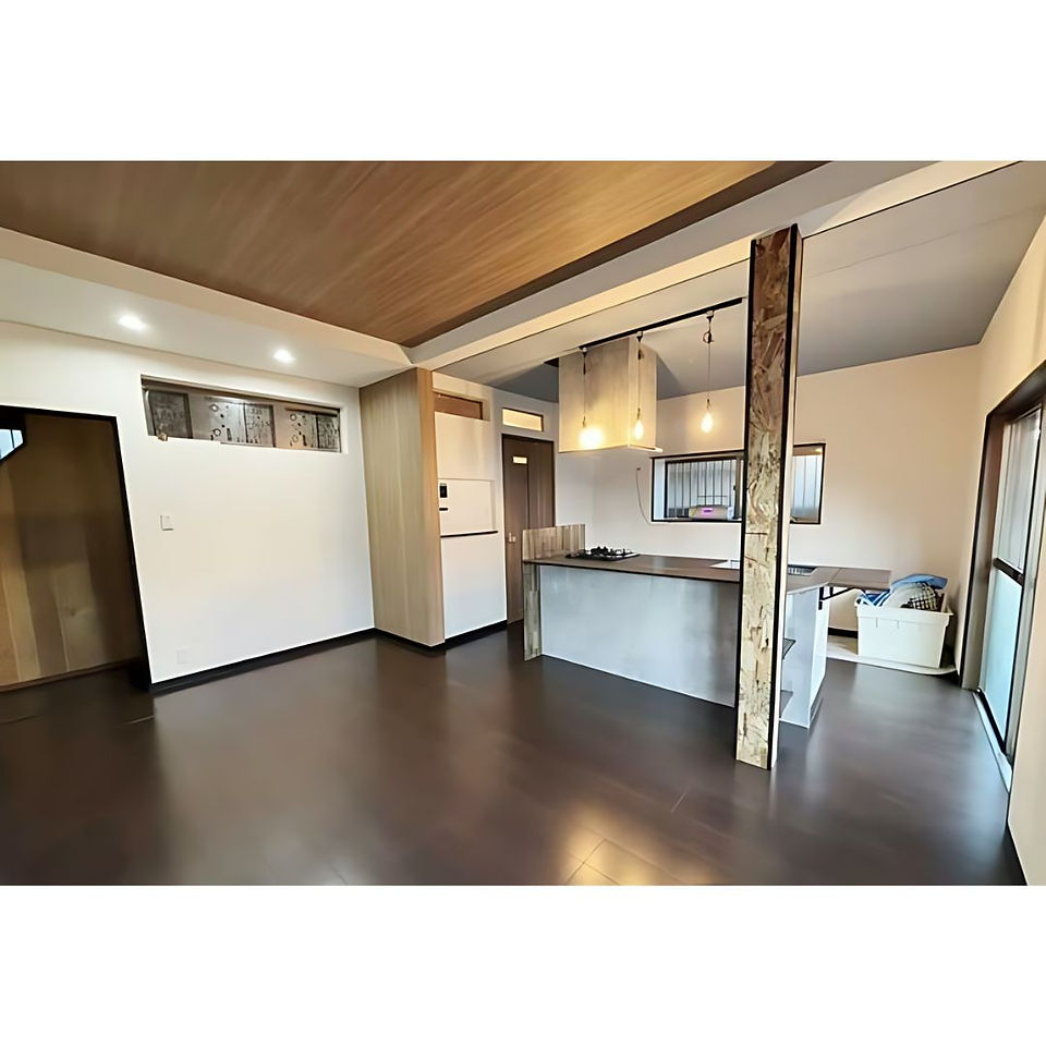 A spacious open-plan living area with a modern kitchen. The kitchen features a concrete island counter with pendant lights overhead. The room has dark wood flooring and a wood-paneled ceiling. Large windows and sliding glass doors provide ample natural light.