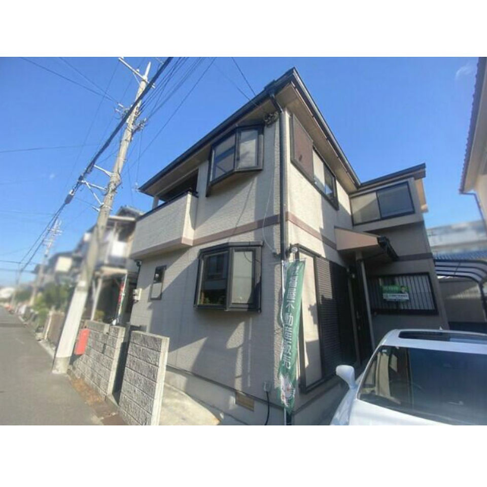 The photo shows a two-story house with a gray facade and a brown roof. The house has a balcony on the second floor and a small front yard with a gate. A car is parked in the driveway. The house is located on a street with other houses visible in the background.