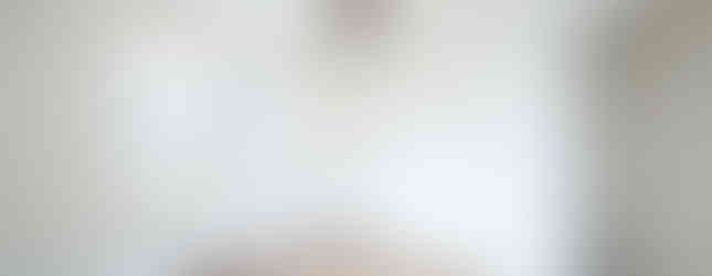 The photo shows a spacious bedroom with white walls and a light wood floor. There are two windows on one wall, letting in natural light. A ceiling fan hangs from the ceiling, and wooden beams add a touch of warmth to the room. Large sliding doors lead to a closet, providing ample storage space. The overall impression is of a clean, bright, and inviting bedroom.