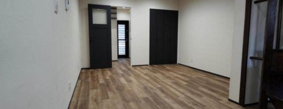 A bright, spacious room with a light wood-look floor and white walls. A pair of dark sliding doors with frosted glass panels are on one side, leading to another room. To the right of the doors is a closet with matching dark doors. Recessed lighting illuminates the space.