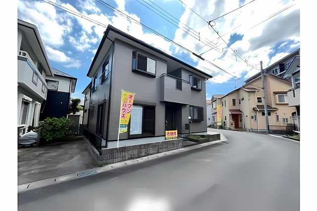 An image of a two-story house with a wooden exterior in a residential neighborhood. The house has a black metal roof and a small balcony. There is a small front yard with a paved walkway and a sign that appears to be advertising the property for sale. The text includes "¥20,900,000" and mentions it is a renovated house.
