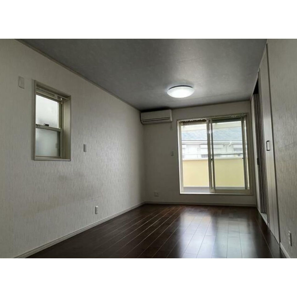 The photo shows a spacious and bright living room with a dark wood floor. There are two windows, one on the side wall and a larger one leading to a balcony, providing plenty of natural light. The walls are painted white, and there is an air conditioning unit mounted on the wall. The overall impression is one of a clean and comfortable space.