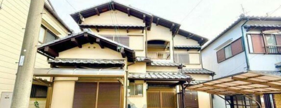 This photo shows the front exterior of a traditional Japanese house. The house is painted light yellow with a dark brown roof and wooden accents. There is a small front porch with a pair of wooden doors. The house is surrounded by other houses and a street.