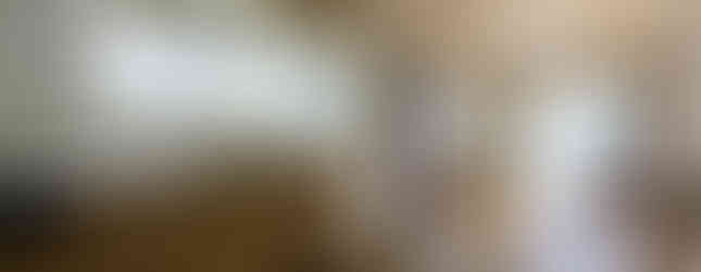 The photograph shows a dated kitchen and dining room area in an apartment. The kitchen features wooden cabinets, a stainless steel sink, and a white refrigerator. A small window is above the sink. The dining area has wooden flooring and a small table with chairs. A balcony door with a view of the outside is visible in the background.