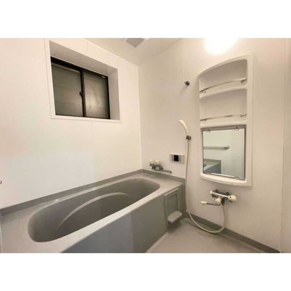 The photo shows a clean and modern bathroom with a bathtub, shower, and sink. The walls are white and the floor is light gray. There is a small window for ventilation and natural light.