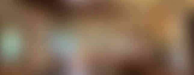 The photo shows a spacious, circular living room with a vaulted wooden ceiling and exposed steel beams. Large windows offer panoramic views of the surrounding greenery. The room is furnished with comfortable seating arrangements, including sofas, armchairs, and a dining table. The overall atmosphere is bright, airy, and inviting.