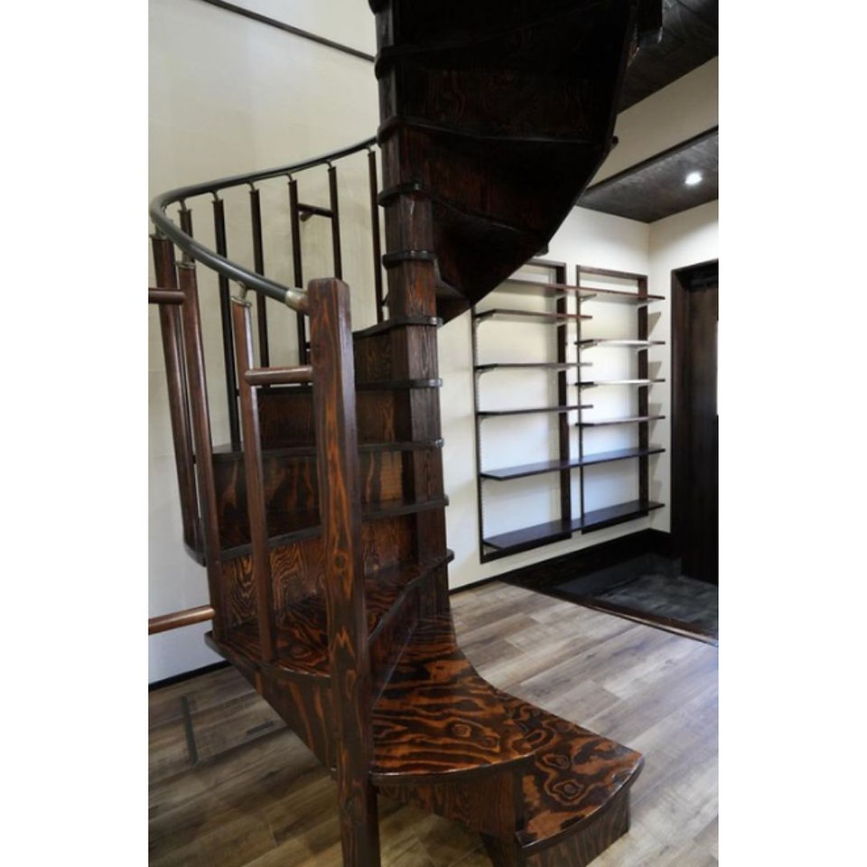 A close-up view of a beautiful wooden spiral staircase with a rich, dark stain. The stairs have open treads and a black metal handrail.  Shelving units are visible along the wall beside the staircase.