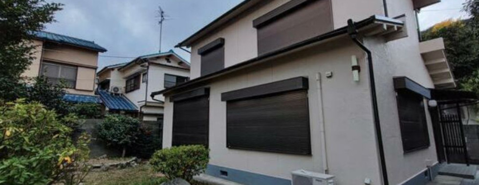 A two-story house with beige walls and black shutters. The house has a small garden in the backyard with a few trees and shrubs. There are other houses visible in the background. The sky is cloudy.
