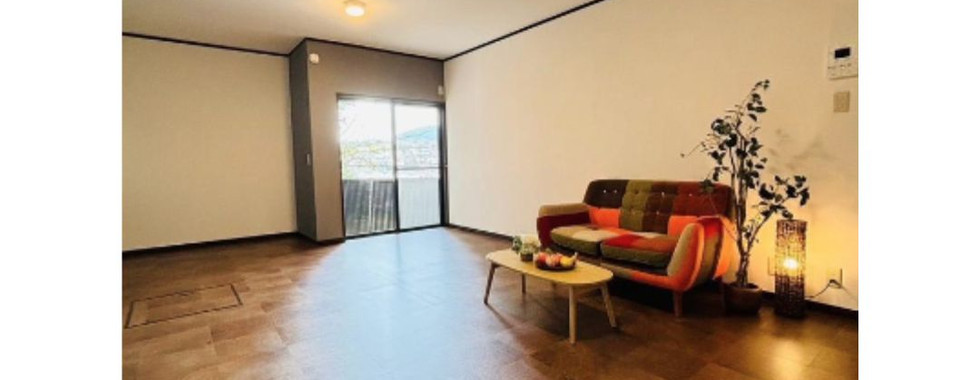 A spacious, well-lit living room with a colorful patchwork sofa, a wooden coffee table, and a potted plant. The room has tiled flooring and recessed lighting, and a sliding glass door leads to a balcony with a view.