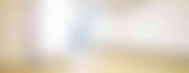 The photograph shows a spacious, empty room with light brown wood flooring and white walls. A doorway with a light-colored door is visible on the left side of the image. In the background, an open doorway leads to a kitchen area, which is partially visible.