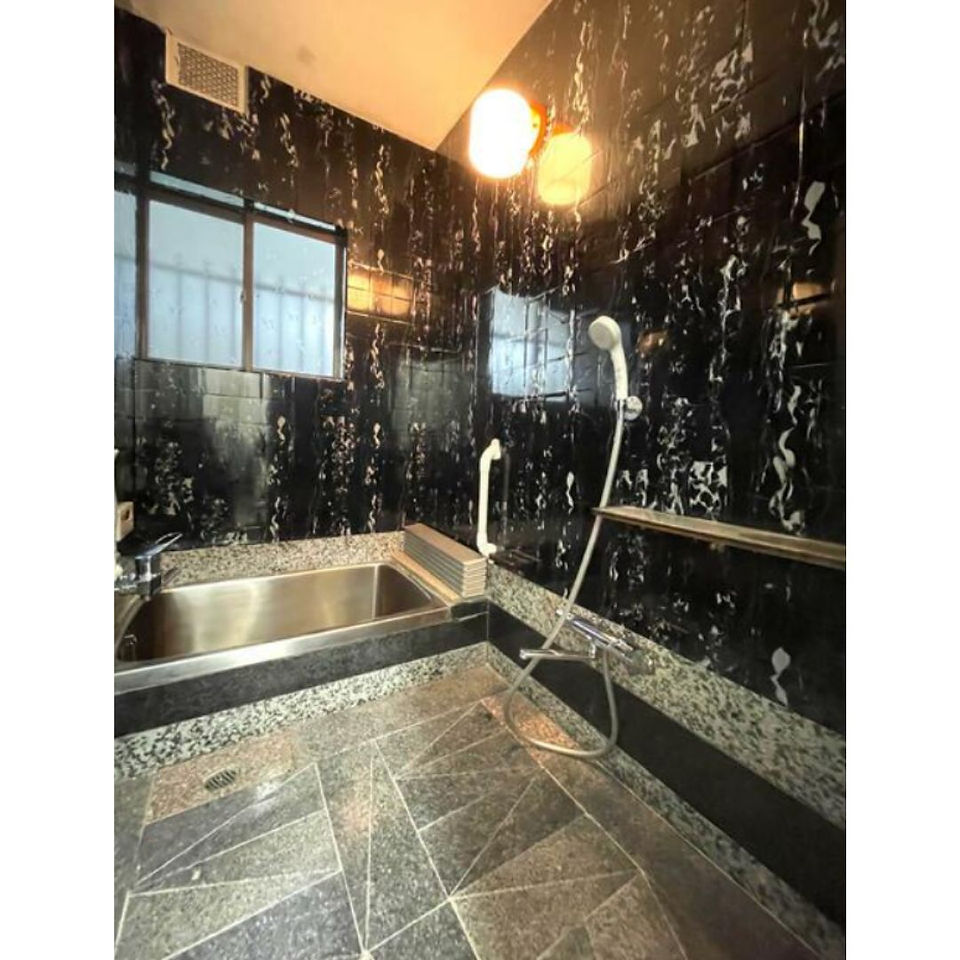 The photograph depicts a bathroom with a luxurious feel. The walls are covered in dark, possibly marble tiles, and the floor has patterned tiles in shades of gray. A stainless steel bathtub is positioned against one wall, with a showerhead and grab bars nearby. The room is illuminated by two pendant lights hanging from the ceiling, creating a warm and inviting atmosphere despite the dark tiles.