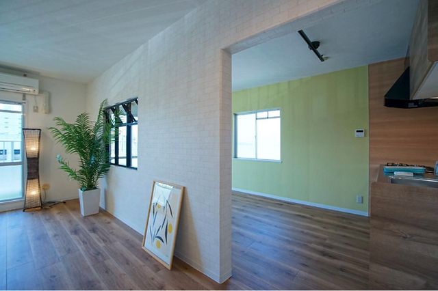 A bright and airy living room with white walls, wooden floors, and large windows. The room is furnished with a sofa, a coffee table, and a potted plant. There is an open doorway leading to the kitchen and dining area.