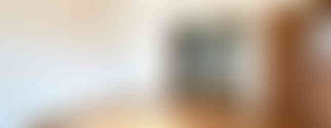 The photo shows a spacious living room with a view into the kitchen. The room has a high ceiling with a wooden trim, white walls, and a wooden floor. Sunlight streams in through a window, illuminating the room.