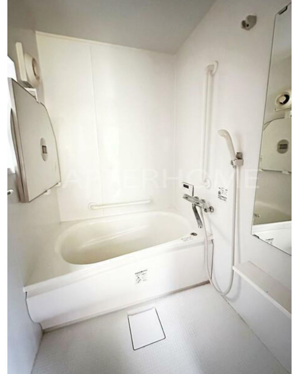 This is a photograph of a clean and functional bathroom in a Japanese style.  The room is compact and features a deep, oval-shaped soaking tub as its focal point. The tub is a light beige color and appears to be made of a smooth, durable material. A chrome faucet with separate hot and cold water handles is mounted on the wall above the tub.  A handheld showerhead is attached to a flexible hose and is hung on the wall.  The walls and floor are a light, neutral color, contributing to the bright and airy feel of the space.  A small, square access panel is visible on the floor, and a larger, hinged panel is on the left wall, possibly for maintenance access to plumbing.  A portion of a mirrored surface is visible on the right side of the image, reflecting part of the room. The overall impression is one of practicality and cleanliness, typical of Japanese bathrooms designed for efficient use of space.  The watermark "AURIHOUSIE" is superimposed on the image.