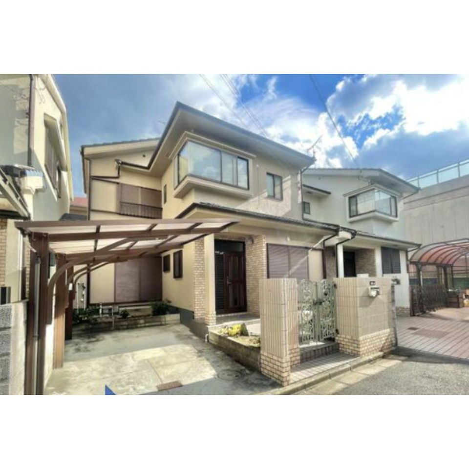 The photo shows a two-story house with a beige facade and a carport. The house has several windows and a small garden in front. The sky is blue and there are some clouds visible. The house is located in a residential neighborhood.
