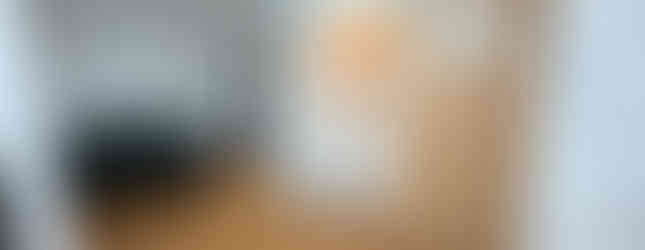 A small apartment living room with a wood floor and white walls. A green couch and coffee table sit on one side, facing a sliding door that opens to a bathroom vanity. A refrigerator is next to the bathroom door, and a weight scale is on the floor.