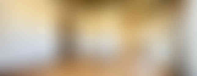 The image shows a spacious living room with a modern and rustic design. The room features a wood-toned ceiling, a concrete-look wall, and a wooden door. The floor is covered in a light wood-colored laminate. A large window provides ample natural light. The room is open to a kitchen area, which is visible in the background.