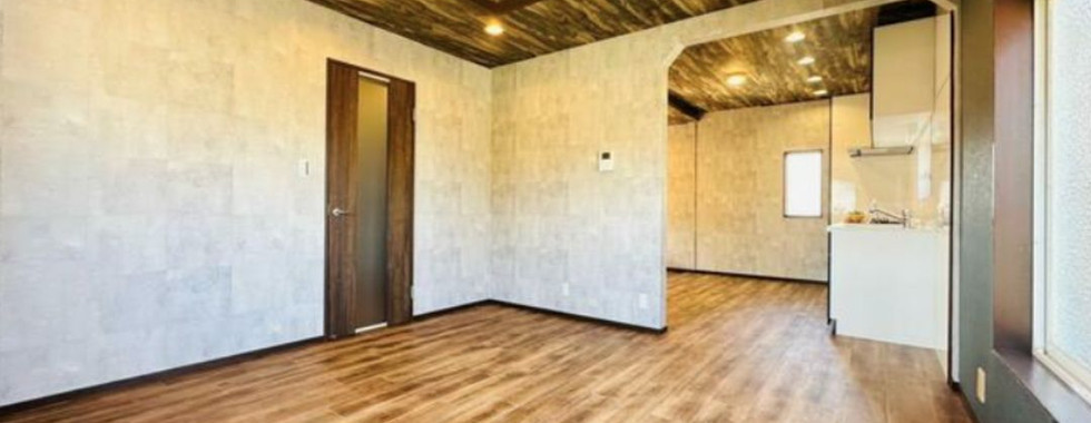 The image shows a spacious living room with a modern and rustic design. The room features a wood-toned ceiling, a concrete-look wall, and a wooden door. The floor is covered in a light wood-colored laminate. A large window provides ample natural light. The room is open to a kitchen area, which is visible in the background.