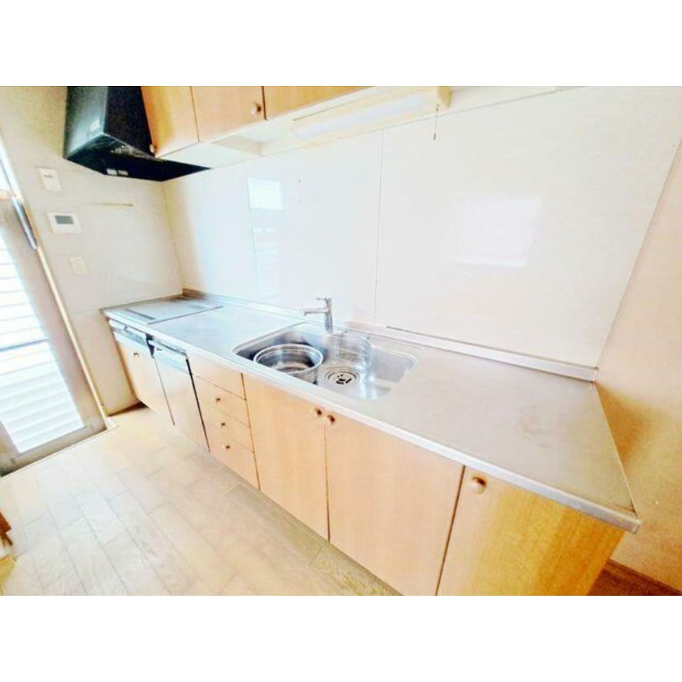 The photo shows a kitchen with light wood cabinets and a stainless steel countertop. The countertop has a sink with a faucet and a built-in stovetop. There is a range hood above the stovetop, and additional cabinets are mounted on the wall above the counter. The floor is covered in light brown tiles.