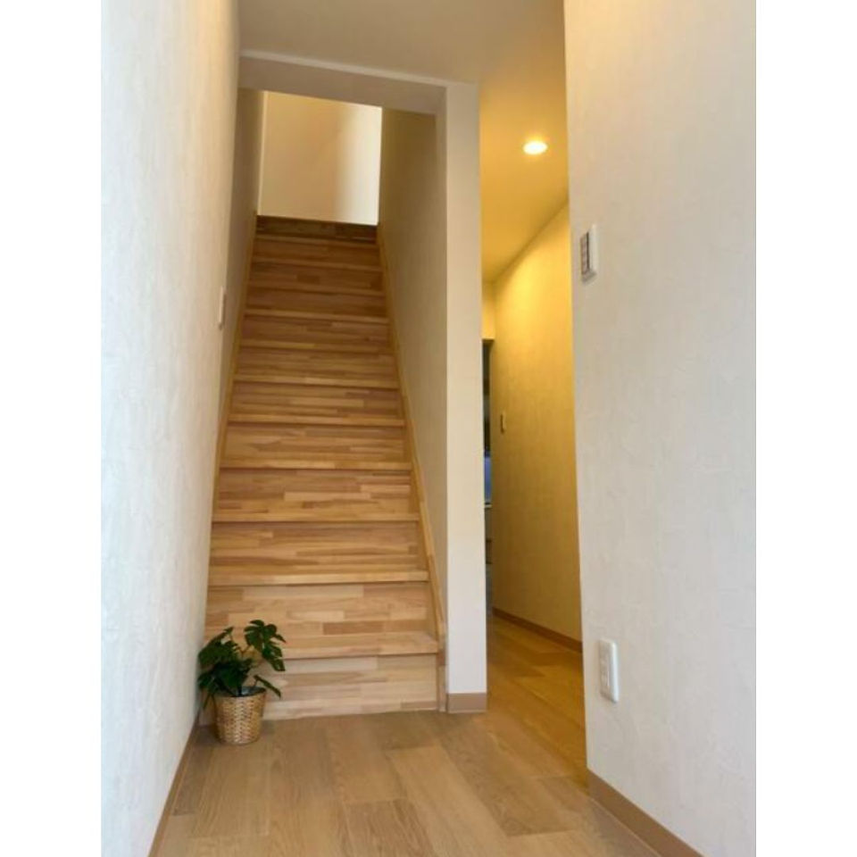 A view from the entrance of a house, showcasing a wooden staircase leading to the upper floor. The walls are painted white and the floor is covered in light wood planks. A potted plant sits at the base of the stairs.