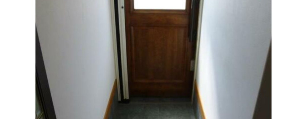 The photograph shows the entryway or genkan of a Japanese house. It features a wooden door with frosted glass panels on the left, leading outside. The floor is covered in gray tiles, with a small step up to a wooden floor further inside the house. A pair of slippers is placed neatly on the wooden floor. The walls are painted white.