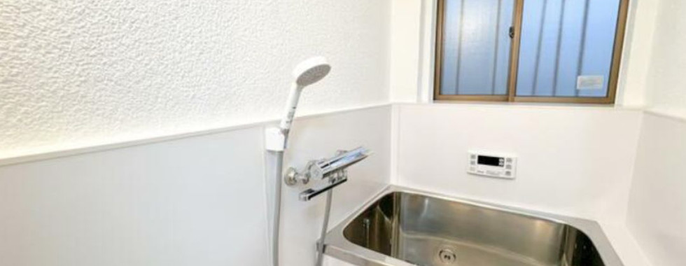 The photo shows a small bathroom with a stainless steel bathtub. There is a shower head and faucet attached to the wall, and a small window provides natural light.