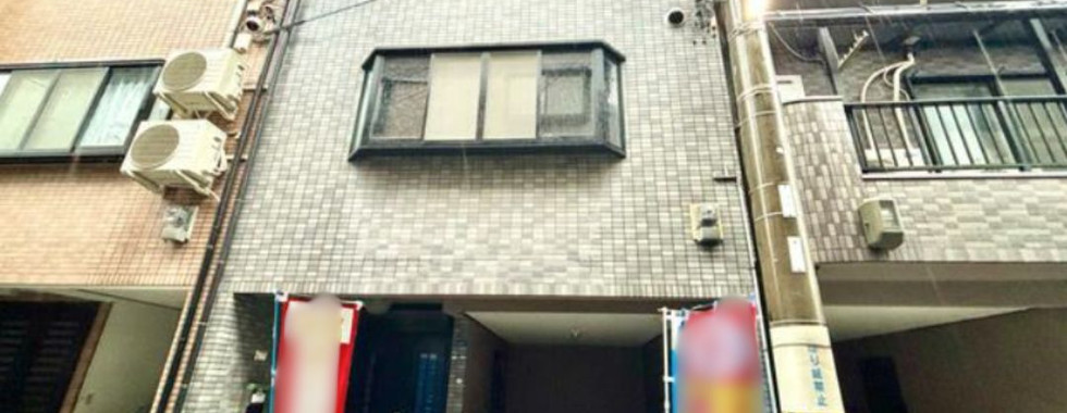 The photo shows a three-story house with a gray facade. The house has several windows and a garage door on the ground floor. There are air conditioning units mounted on the side of the building. The house is located on a street with other houses nearby.